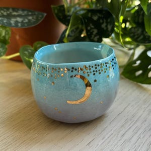 Może przedstawiać: Mały, zaokrąglony kubek ceramiczny z gradientem kolorów przechodzącym od jasnego błękitu u góry do delikatnego lawendowego u podstawy. Zewnętrzna strona ozdobiona jest metalicznym złotym półksiężycem i rozproszonymi złotymi kropkami.