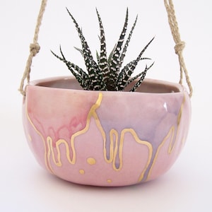 May include: A hanging planter with a succulent plant. The ceramic pot is a blend of pink and lavender hues, with gold accents dripping down the sides. The plant has dark green leaves with white spots, and the pot is suspended by a natural rope.