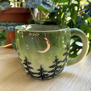 May include: A light green ceramic mug with a hand-painted forest of dark pine trees around the base. The upper portion features a metallic gold crescent moon and a series of small gold dots resembling stars against the green glaze.