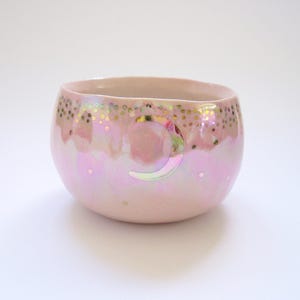Könnte beinhalten: Eine kleine, abgerundete Keramikschale mit glänzendem, irisierendem rosa Finish. Die Außenseite zeigt ein goldenes Halbmond-Motiv nahe dem Rand, umgeben von einem Muster aus kleinen goldenen Punkten und dezenten, verstreuten weißen Akzenten.