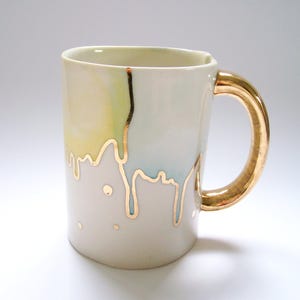Könnte beinhalten: Eine Keramik-Tasse mit goldenem Henkel und goldenem Tropfendesign. Die Tasse hat eine cremefarbene Basis mit pastellgelben und blauen Akzenten. Die goldenen Akzente erzeugen einen einzigartigen, künstlerischen Look. Perfekt für Kaffee, Tee oder jedes Getränk.