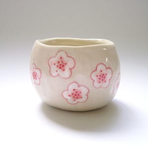 May include: A small, off-white ceramic cup with a rounded shape. The cup is decorated with pink cherry blossom designs. Each blossom has a pink center with small dots. The cup has a smooth, glossy finish.