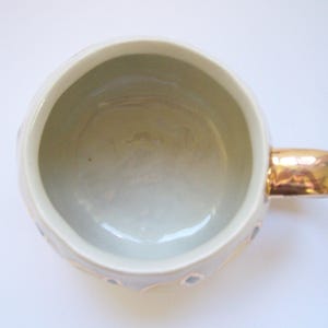 Może przedstawiać: Ceramiczny kubek widziany z g&oacute;ry, z gładkim, jasnym wnętrzem i odblaskowym uchwytem w kolorze złota. Zewnętrzna powierzchnia posiada subtelny, fasetowany wz&oacute;r geometryczny z małymi, przygaszonymi niebieskimi akcentami.