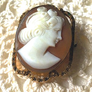 May include: An oval-shaped cameo brooch featuring a white carved profile of a person with an elaborate hairstyle set against a light brown background. The cameo is encased in a decorative gold-toned metal frame with a textured, scalloped edge.