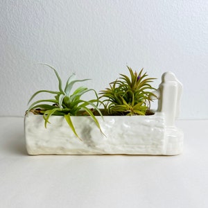 May include: White ceramic planter shaped like a log with two air plants inside. The planter has a textured surface and a small, raised figure on the side.
