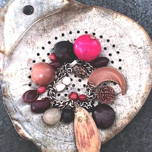 May include: A charm bracelet featuring a variety of natural elements including dark brown seeds, a bright pink sphere, a crescent-shaped wooden piece, a textured brown bead, and a large elongated seed pod with a small metal cap, all on a metal chain.