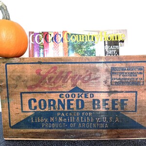 May include: An orange pumpkin rests on a weathered wooden box labeled "Libby's Cooked Corned Beef." The box has blue and red lettering and is packed for Libby, McNeill & Libby, U.S.A. Magazines are stacked behind the pumpkin.