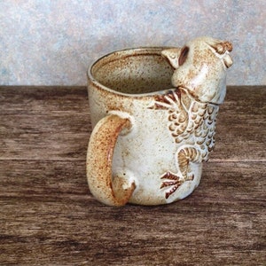 May include: A ceramic mug with a brown and white speckled glaze. The mug has a handle and a bird-shaped spout. The bird is brown and white with a speckled glaze.