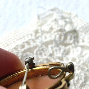 May include: A gold-toned metal brooch featuring a pin mechanism and a small loop on the back. The metal frame is stamped with the text 1/20 12K G.F. AMCO. The brooch has a smooth, light-colored, oval-shaped center insert.