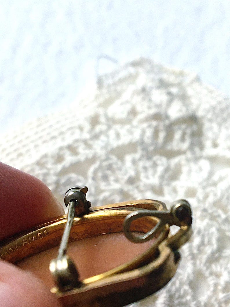 May include: A gold-toned metal brooch featuring a pin mechanism and a small loop on the back. The metal frame is stamped with the text 1/20 12K G.F. AMCO. The brooch has a smooth, light-colored, oval-shaped center insert.