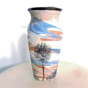 May include: A ceramic vase with a flared rim and a dark brown top edge. The body features a marbled pattern of soft blue, peach, and white swirls. A detailed black ink illustration of a tall, leafless tree stands prominently in the center.
