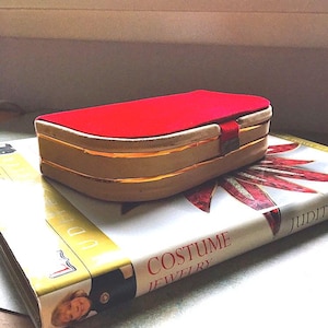 K&ouml;nnte beinhalten: Eine rechteckige, vintage Schmuckschatulle mit rotem Samtdeckel und goldenen Zierleisten. Die Schatulle liegt auf einem Buch mit dem Titel "Costume Jewelry". Das Buch hat einen wei&szlig;en Einband mit roten und goldenen Akzenten.