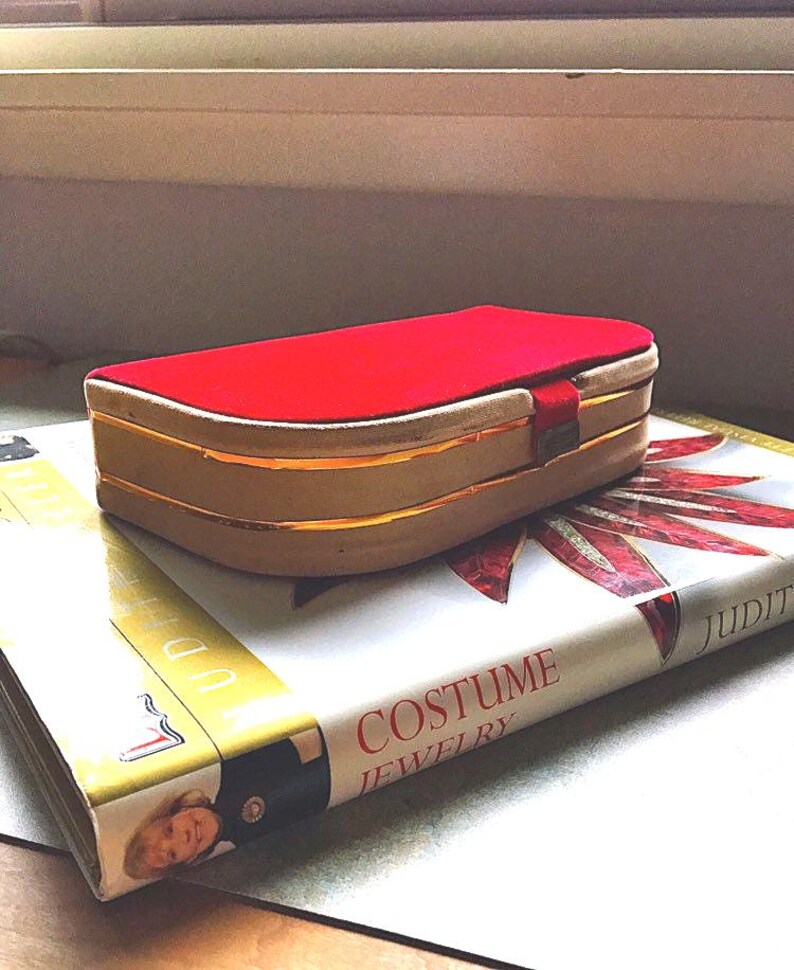 K&ouml;nnte beinhalten: Eine rechteckige, vintage Schmuckschatulle mit rotem Samtdeckel und goldenen Zierleisten. Die Schatulle liegt auf einem Buch mit dem Titel "Costume Jewelry". Das Buch hat einen wei&szlig;en Einband mit roten und goldenen Akzenten.