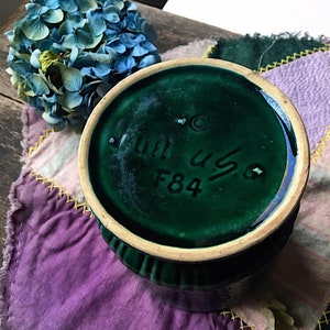 May include: Green ceramic vase with a textured rim and a white base. The bottom of the vase has the text "Juli us 6 F84" in black.