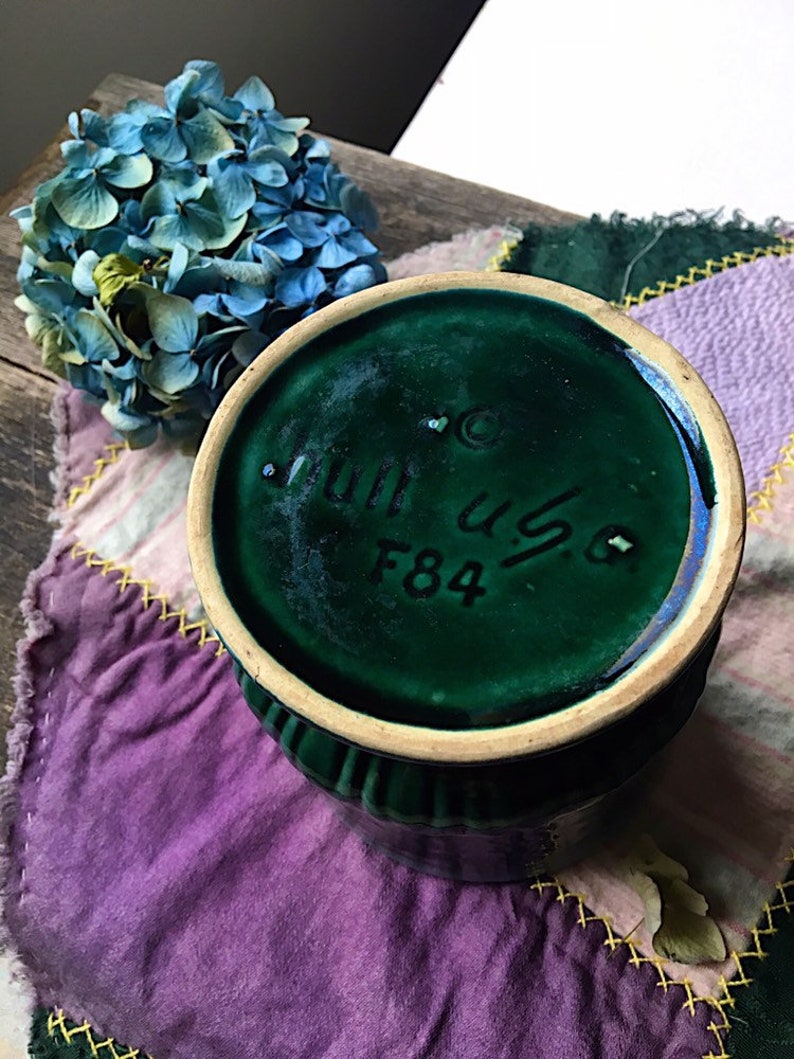 May include: Green ceramic vase with a textured rim and a white base. The bottom of the vase has the text "Juli us 6 F84" in black.