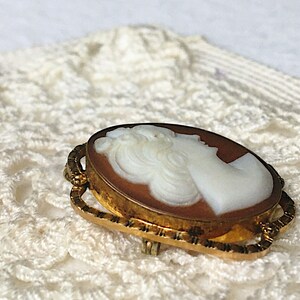 May include: An oval-shaped brooch featuring a white carved profile of a person set against a brown background. The cameo is encased in a textured gold-toned metal frame with an intricate, openwork border design and a pin mechanism on the back.