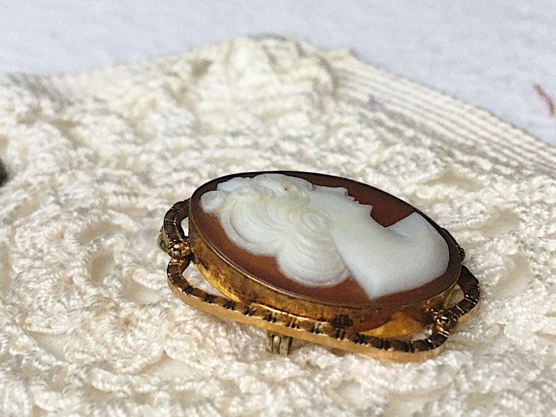 May include: An oval-shaped brooch featuring a white carved profile of a person set against a brown background. The cameo is encased in a textured gold-toned metal frame with an intricate, openwork border design and a pin mechanism on the back.