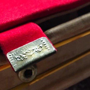K&ouml;nnte beinhalten: Nahaufnahme eines roten Samtstoffs mit einem Metallverschluss. Der Verschluss hat ein eingraviertes Design und ist an einer Holzstruktur befestigt. Das Bild hebt die Textur und die Details der Materialien hervor.