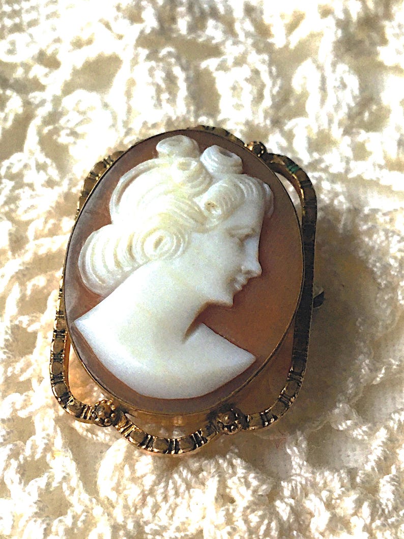 May include: An oval-shaped cameo brooch featuring a white carved profile of a person with an elaborate hairstyle set against a light brown background. The cameo is encased in a decorative gold-toned metal frame with a textured, scalloped edge.