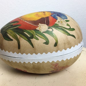 May include: A vintage Easter egg-shaped box with a white scalloped edge. The box is decorated with a floral design featuring yellow, red, blue, and green colors. The design includes a bird, flowers, and leaves.