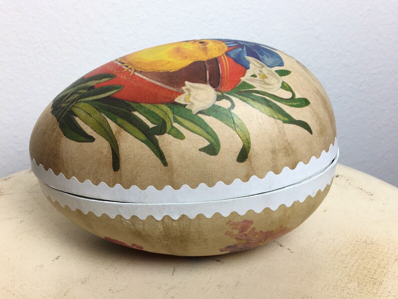 May include: A vintage Easter egg-shaped box with a white scalloped edge. The box is decorated with a floral design featuring yellow, red, blue, and green colors. The design includes a bird, flowers, and leaves.