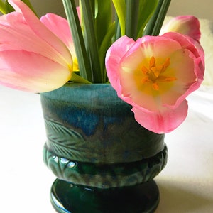 May include: A green ceramic vase with a leafy design holds pink tulips. The vase has a wide base and a narrow neck.