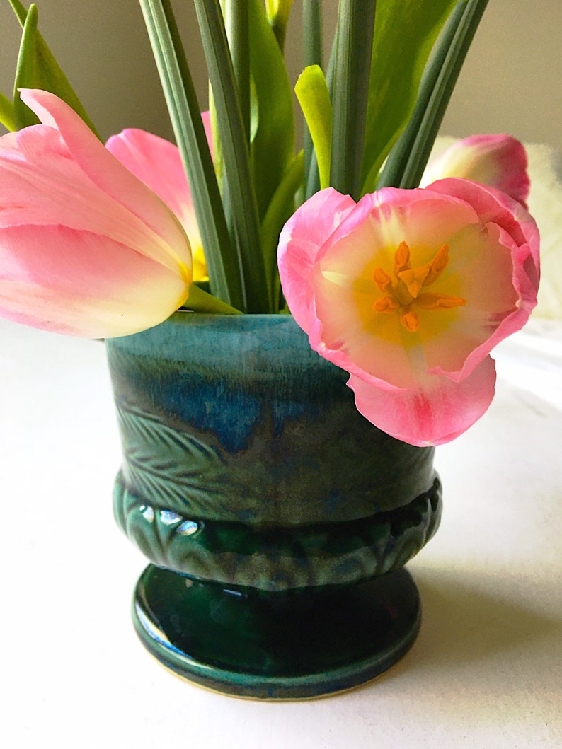May include: A green ceramic vase with a leafy design holds pink tulips. The vase has a wide base and a narrow neck.