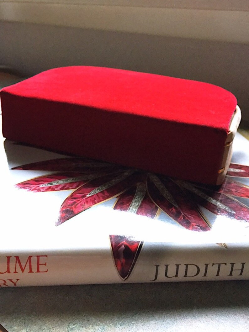 K&ouml;nnte beinhalten: Eine rechteckige, rote Samt-Schmuckschatulle mit goldenem Rand liegt auf einem Buch mit dem Titel "Judith". Die geschlossene Schatulle enth&auml;lt wahrscheinlich Schmuck oder andere kleine Wertgegenst&auml;nde. Das Buchcover zeigt ein rot-wei&szlig;es Design.