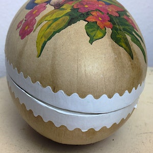 May include: A vintage Easter egg-shaped container made of paper mache with a white scalloped edge. The egg is decorated with a colorful floral design and a bird.