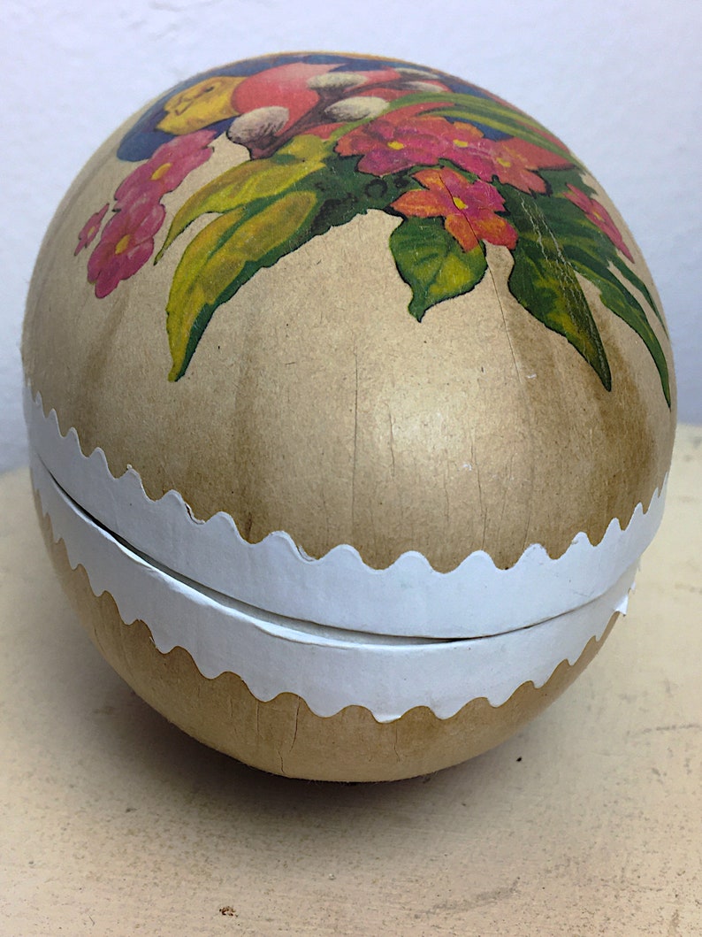 May include: A vintage Easter egg-shaped container made of paper mache with a white scalloped edge. The egg is decorated with a colorful floral design and a bird.