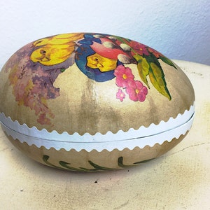 May include: A vintage Easter egg-shaped box with a white scalloped edge. The box is decorated with a colorful floral design featuring yellow chicks, pink flowers, and green leaves on a beige background.