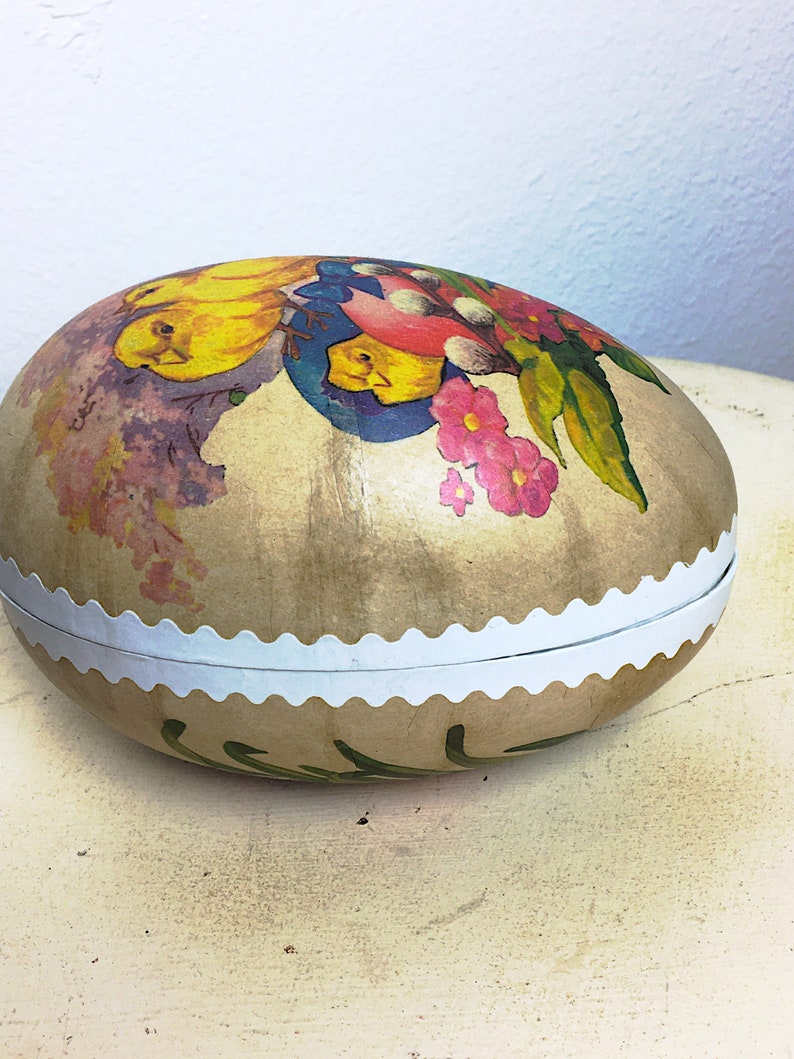 May include: A vintage Easter egg-shaped box with a white scalloped edge. The box is decorated with a colorful floral design featuring yellow chicks, pink flowers, and green leaves on a beige background.
