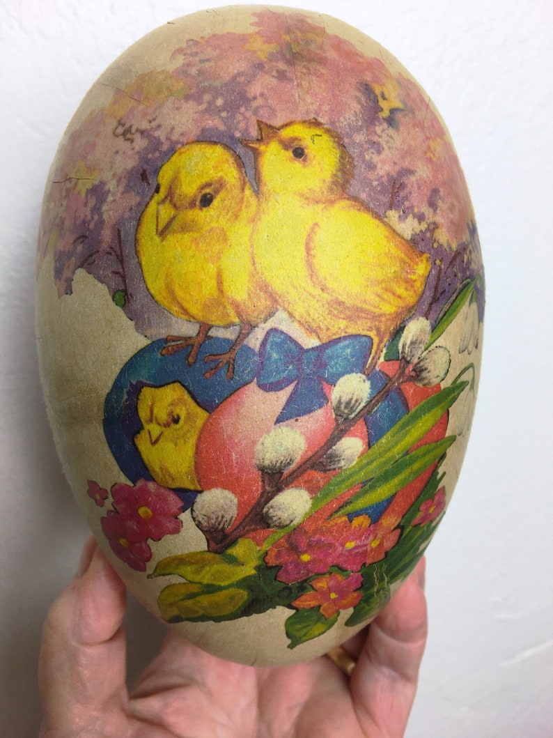 May include: A vintage Easter egg made of paper mache with a colorful illustration of two yellow chicks, a pink heart, and spring flowers. The egg is decorated with a blue ribbon and has a textured surface.