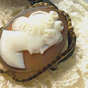 May include: An oval cameo brooch featuring a white carved profile of a person with curled hair set against a brown background. The cameo is encased in a decorative gold-toned metal frame with intricate textured detailing along the edges.