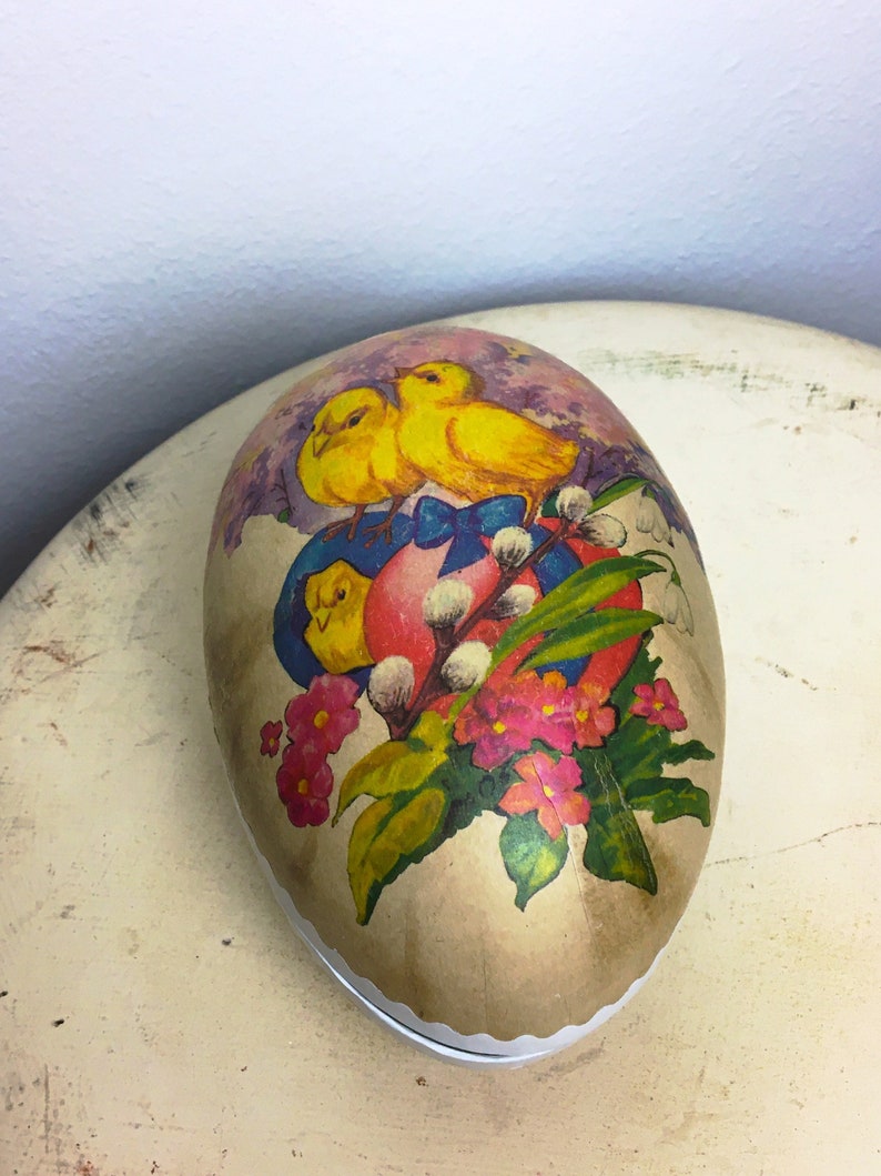 May include: A vintage Easter egg-shaped container with a paper mache exterior. The container is decorated with a colorful illustration of two yellow chicks, a pink egg, a blue ribbon, and white flowers.