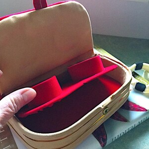 K&ouml;nnte beinhalten: Ge&ouml;ffnete, hellfarbene Schmuckschatulle mit rotem Innenraum und roter Trennwand. Die Schatulle hat einen Klappdeckel und einen Metallverschluss. Die Schatulle liegt auf einem Buch.