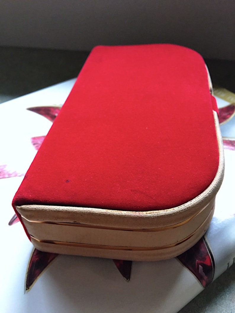 K&ouml;nnte beinhalten: Eine rechteckige rote Samtbox mit abgerundeter Oberseite. Die Box hat einen hellbeigen Rand mit einer d&uuml;nnen goldenen Zierleiste. Die Box liegt auf einer gemusterten Oberfl&auml;che.