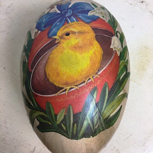 May include: A vintage Easter egg made of paper mache with a yellow chick inside a red basket. The egg is decorated with a blue bow, white flowers, and green leaves.