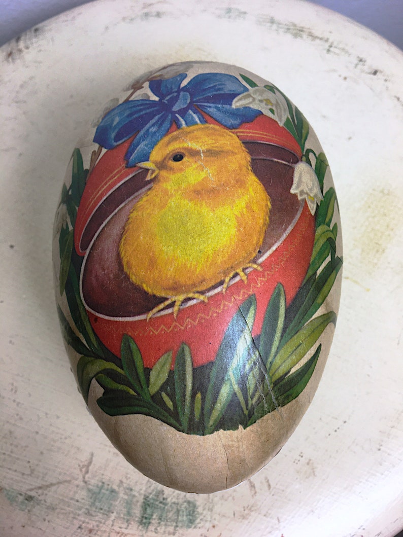 May include: A vintage Easter egg made of paper mache with a yellow chick inside a red basket. The egg is decorated with a blue bow, white flowers, and green leaves.