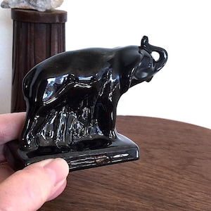 May include: A glossy black ceramic elephant figurine. The elephant has its trunk raised and is standing on a rectangular base. The figurine is detailed with visible wrinkles and folds. The background includes a wooden surface and a dark wooden container.