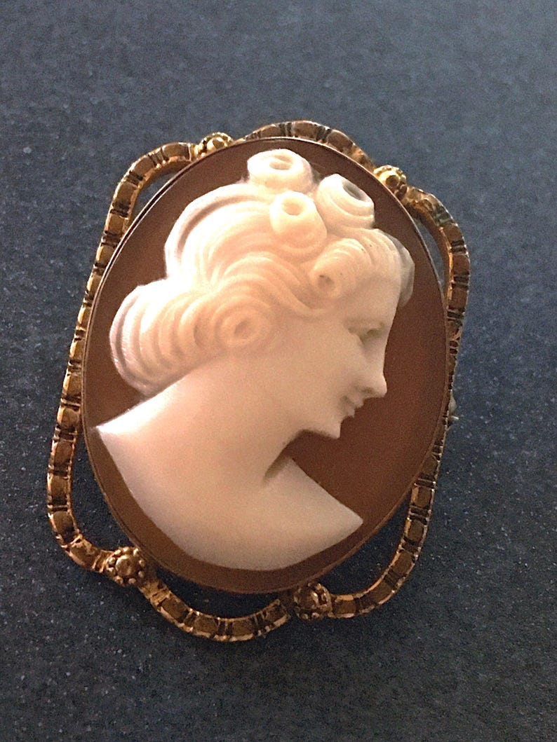 May include: An oval cameo brooch featuring a white carved profile of a person with curled hair set against a brown background. The cameo is framed by an ornate, textured gold-tone metal border with small decorative accents at the top and bottom.