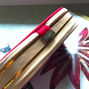 K&ouml;nnte beinhalten: Eine goldfarbene Clutch mit einem roten Samtband und einem silbernen Verschluss. Die Clutch ist ge&ouml;ffnet und zeigt ein goldenes Innenfutter. Ein dekoratives rot-wei&szlig;es Blumendesign ist im Hintergrund zu sehen.