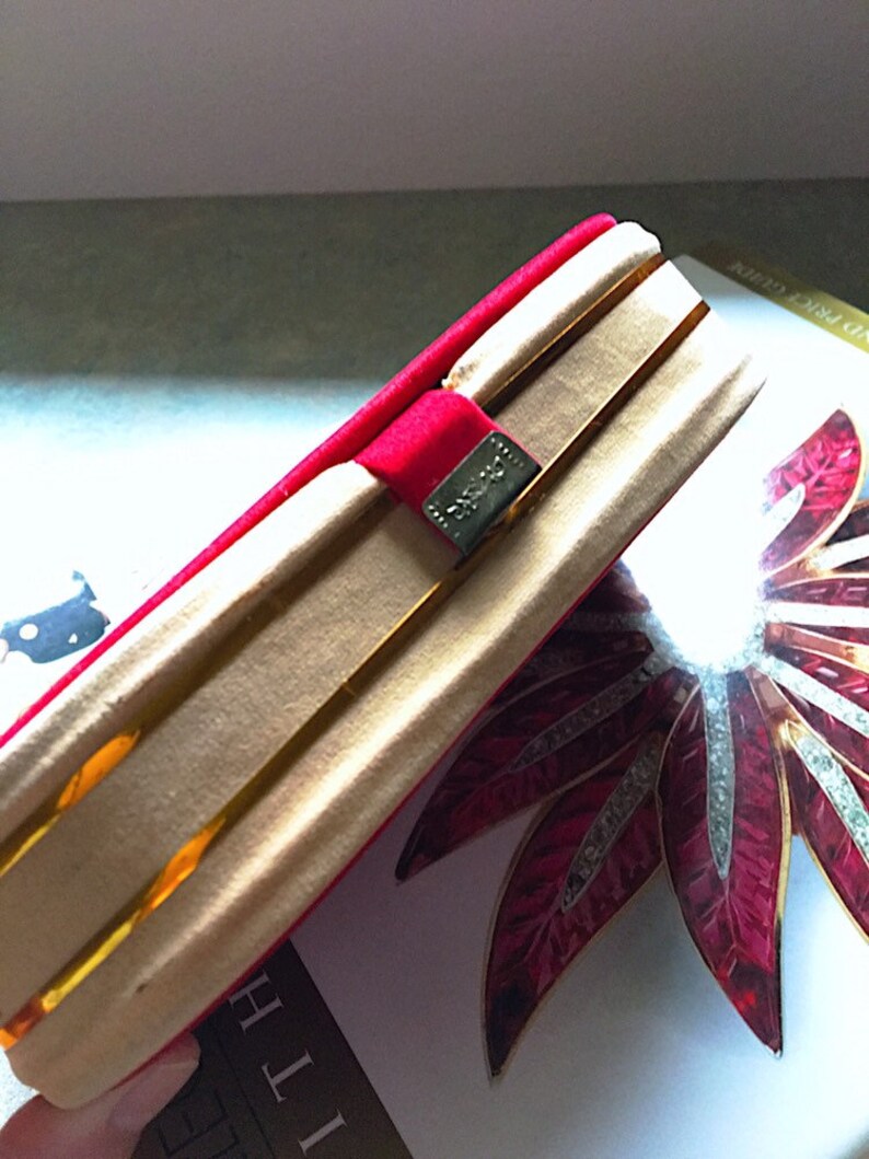 K&ouml;nnte beinhalten: Eine goldfarbene Clutch mit einem roten Samtband und einem silbernen Verschluss. Die Clutch ist ge&ouml;ffnet und zeigt ein goldenes Innenfutter. Ein dekoratives rot-wei&szlig;es Blumendesign ist im Hintergrund zu sehen.