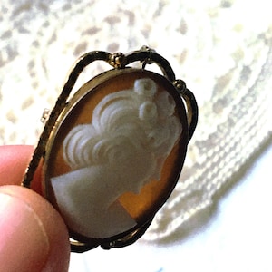 May include: An oval-shaped brooch featuring a carved white profile of a person with an updo hairstyle set against a translucent amber-colored background. The cameo is encased in a textured gold-toned metal frame with decorative arches.