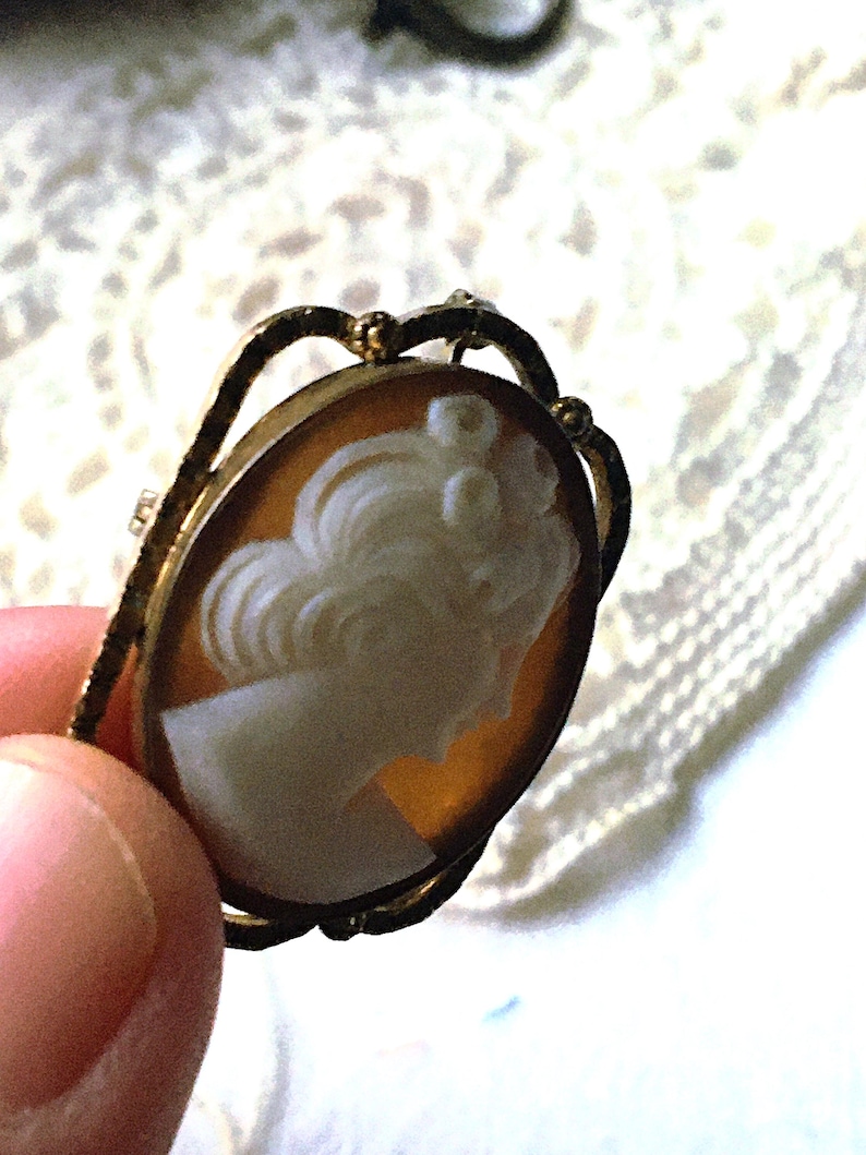 May include: An oval-shaped brooch featuring a carved white profile of a person with an updo hairstyle set against a translucent amber-colored background. The cameo is encased in a textured gold-toned metal frame with decorative arches.