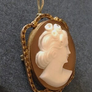 May include: An oval-shaped cameo brooch and pendant featuring a white carved profile of a person with curled hair set against a light brown background. The piece is encased in a textured gold-toned metal frame with a small loop at the top.