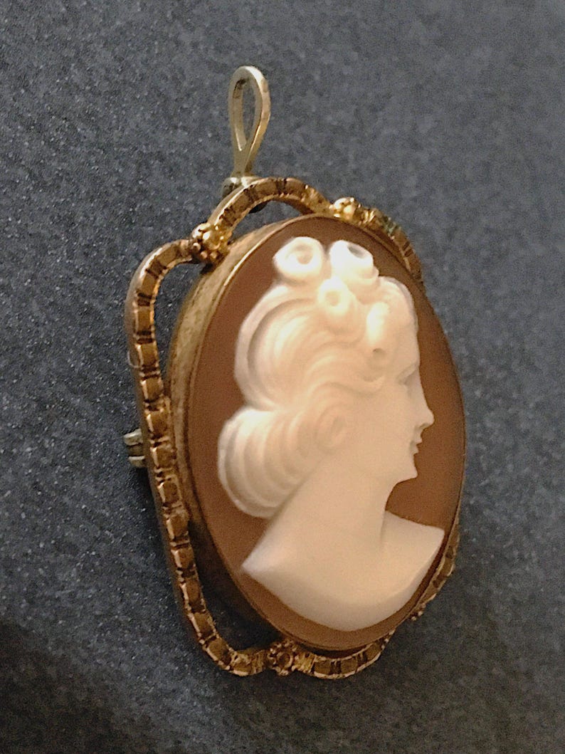 May include: An oval-shaped cameo brooch and pendant featuring a white carved profile of a person with curled hair set against a light brown background. The piece is encased in a textured gold-toned metal frame with a small loop at the top.