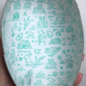 May include: A white Easter egg-shaped container with a green and white design. The container is decorated with images of bunnies, birds, flowers, and eggs. The text "Echt Erzgebirge" and "Made in Germany" are printed on the container.