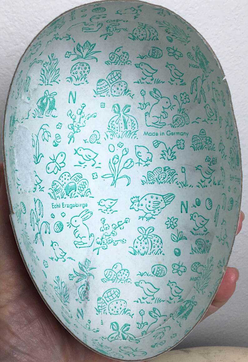 May include: A white Easter egg-shaped container with a green and white design. The container is decorated with images of bunnies, birds, flowers, and eggs. The text "Echt Erzgebirge" and "Made in Germany" are printed on the container.