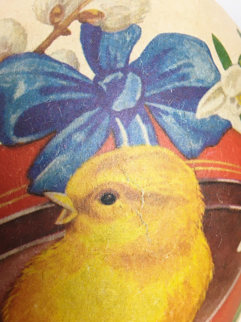 May include: A vintage Easter egg with a yellow chick and a blue bow. The egg is decorated with white flowers and a red and white striped band.